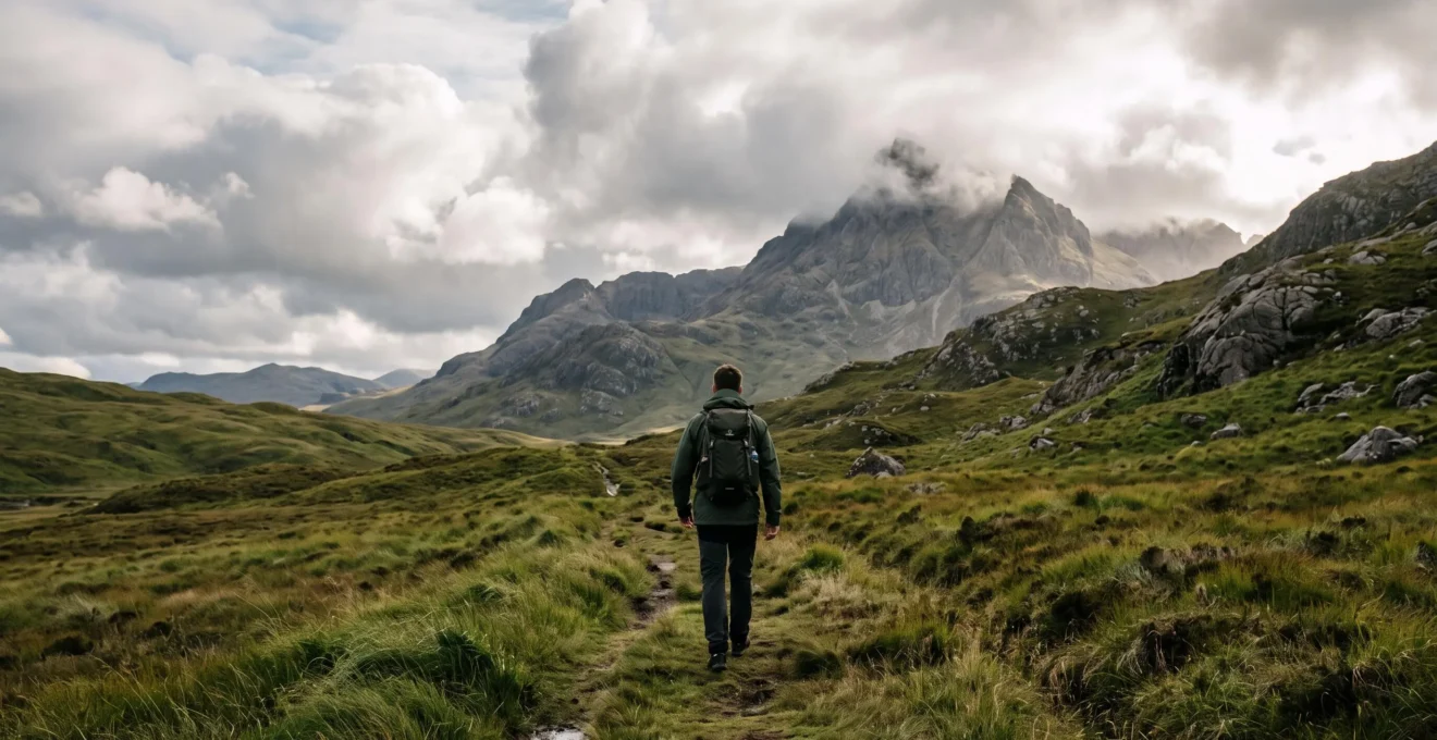 A hiker transitioning from a gentle green lowland trail toward a rugged, cloud-capped UK mountain ridge