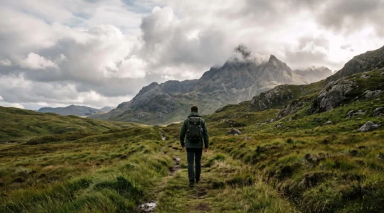 A hiker transitioning from a gentle green lowland trail toward a rugged, cloud-capped UK mountain ridge