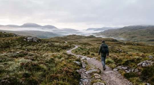A lone hiker walking respectfully along a marked stone path through a misty UK upland national park, surrounded by untouched moorland.